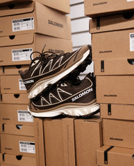 Salomon trail sneaker bruin en wit met veters, naast gestapelde schoenendozen.